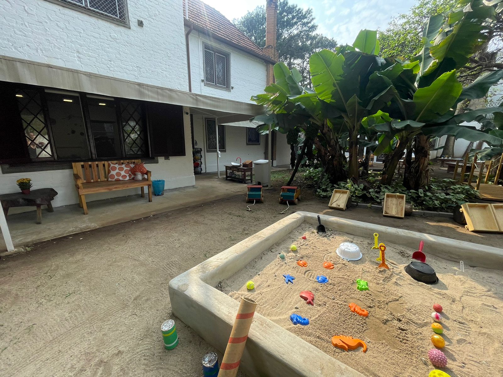 Bananeiras, pia de madeira e regadores no caminho do quintal